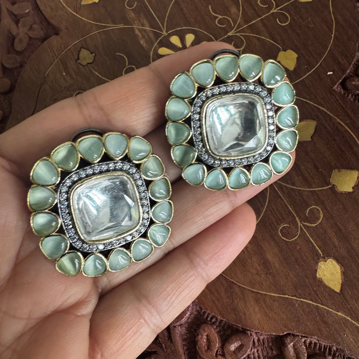 Decorative earrings held by a hand on a wooden surface with intricate carvings