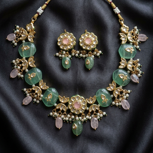 An ornate gold-toned jewelry set featuring a statement necklace and matching earrings, embellished with green faceted beads, pink stones, and small pearl-like accents, displayed on a dark satin fabric background.